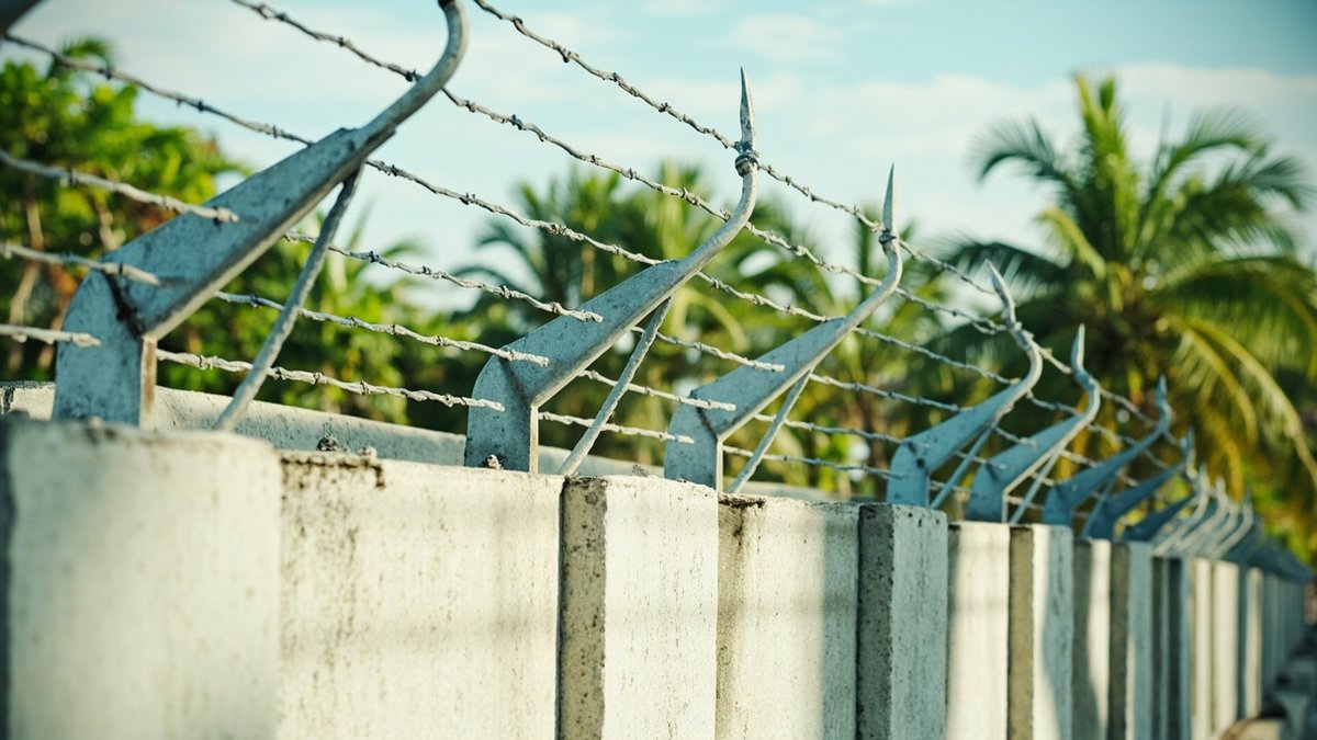 Barbed wire strands on angled steel brackets along the top of a plastered boundary wall