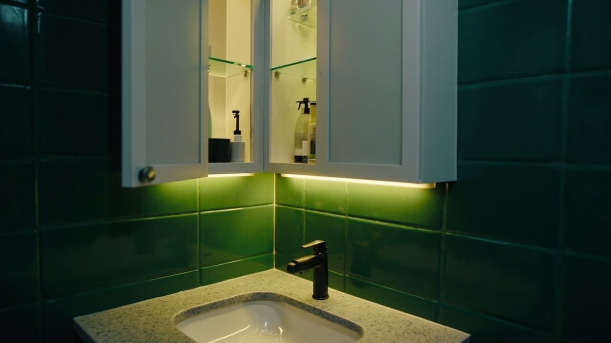Three-door bathroom mirror cabinet with one door open over a basin, lit by an LED strip below