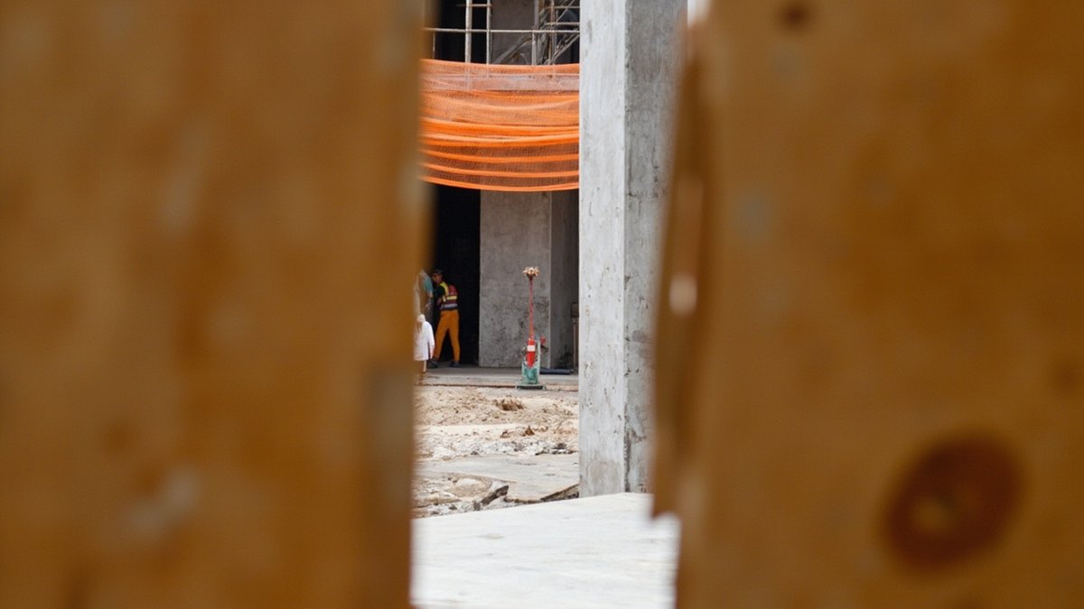A gap in a hoarding board reveals a raw building site, representing a deed of assignment before strata title issues.