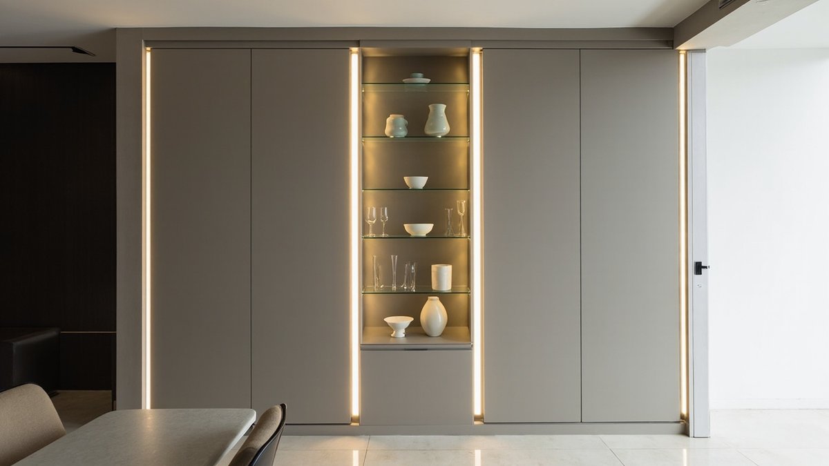 Illustrative concept design: warm-grey cabinet wall with a central lit display bay of glass shelves and celadon vases beside a dining table