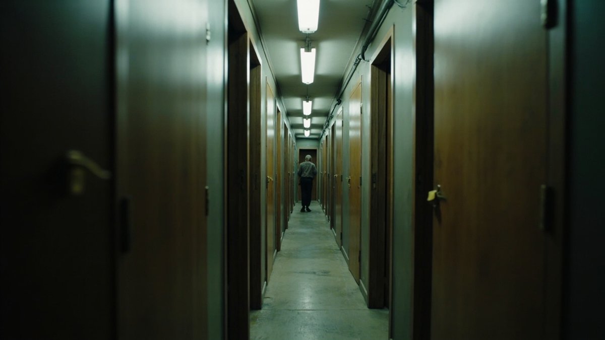 Narrow corridor lined with closed wooden room doors under fluorescent lights, a person walking away in the distance
