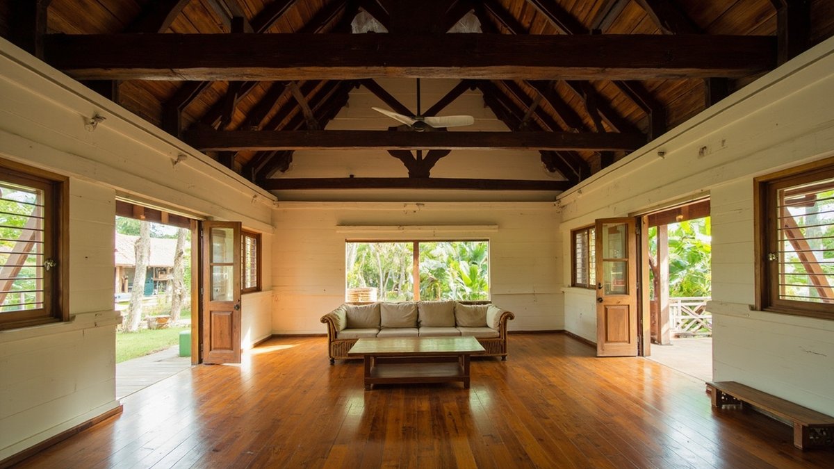 Illustrative concept design: cream plank-walled living hall under an exposed timber roof with a ceiling fan, a rattan-framed sofa and timber louvred windows