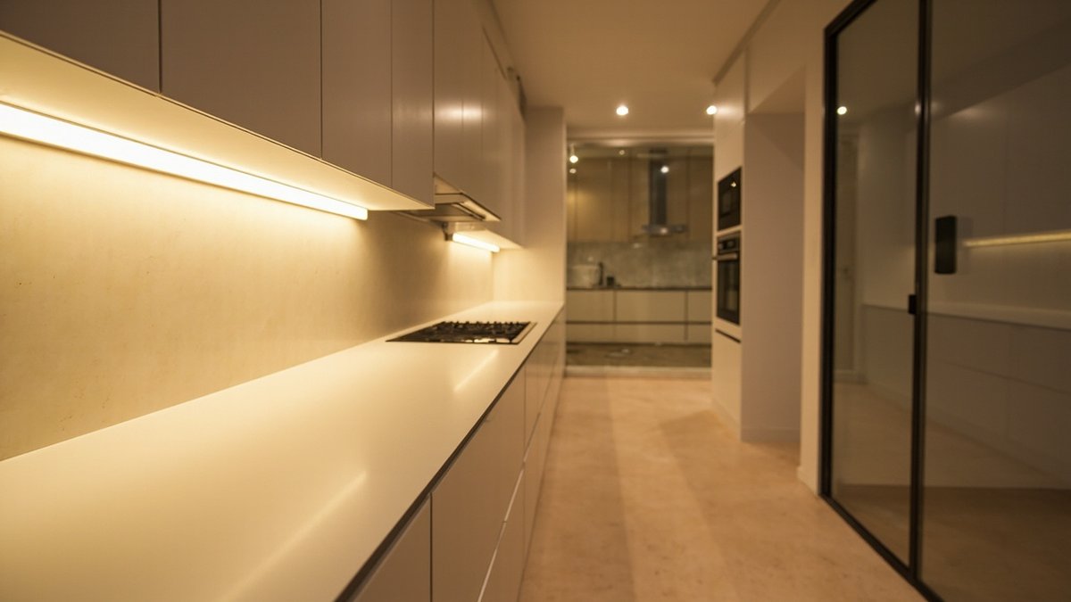 Ilustrasi konsep reka bentuk: dapur galley pada waktu malam dengan jalur led suam di bawah kabinet dinding matt menyinari splashback rupa batu pucat dan table top putih kosong
