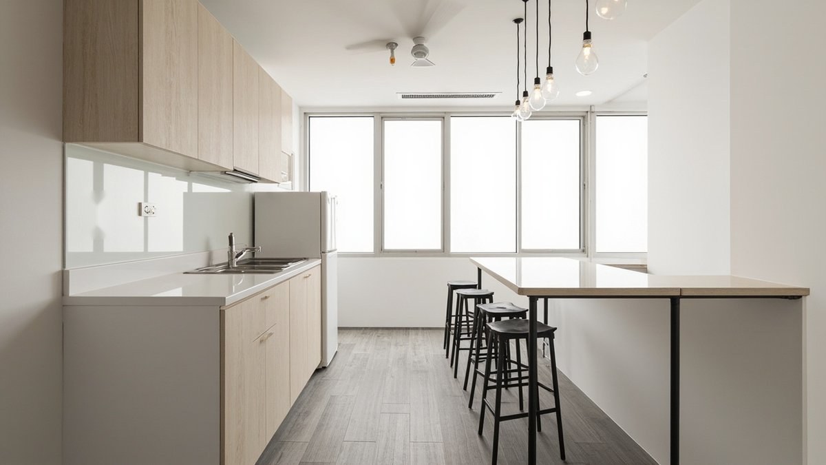 Illustrative concept design: galley office pantry with light oak cabinets, a white worktop and sink, a plain fridge and a high table with black stools by the windows