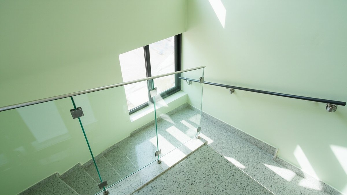 Ilustrasi konsep reka bentuk: balustrade kaca pada tangga rupa terrazzo yang membelok di landing, dengan susur tangan hitam di dinding