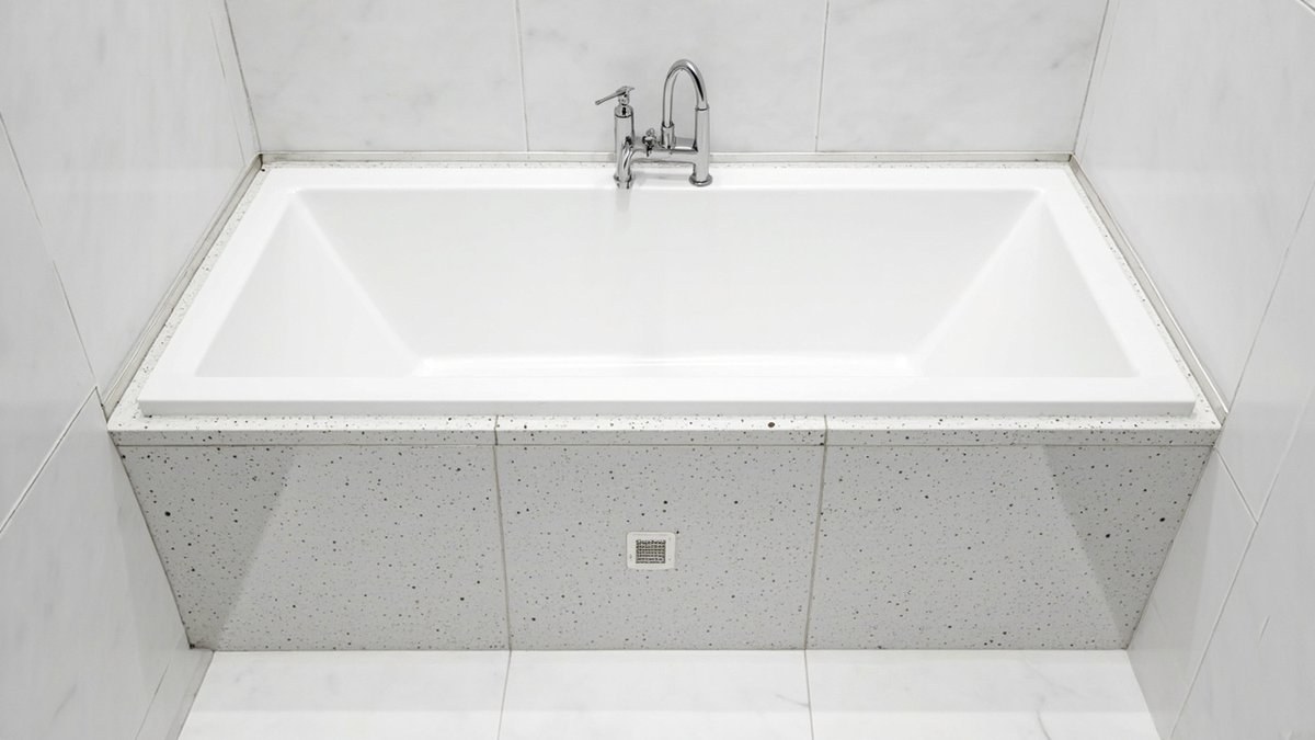 Illustrative concept design: built-in bathtub with a terrazzo-look tiled apron and a small square access hatch in a white bathroom